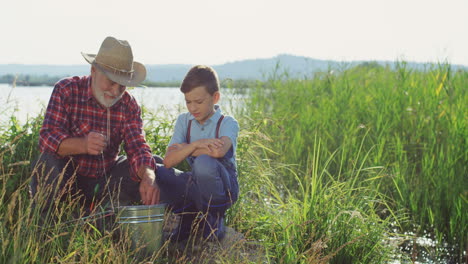 Senior-fisherman-in-a-hat-with-his-grandson-sitting-on-the-shore-of-the-lake-and-putting-a-fish-in-the-backet