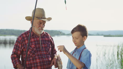 Grandfather-standing-with-his-teen-grandson-at-the-lake-shore,-talking-and-holding-a-fish-on-the-rod
