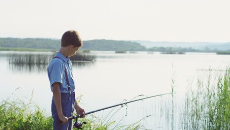 Teenager-Junge,-Der-Morgens-An-Einem-Sommertag-Am-Malerischen-Seeufer-Mit-Einer-Großen-Rute-Fischt