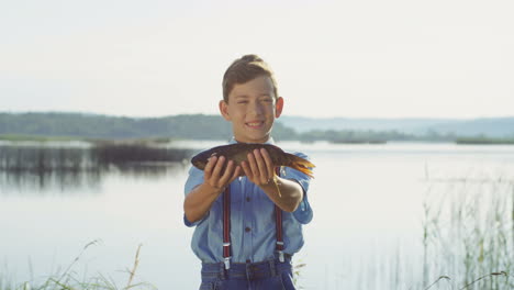 Portrait-of-a-cute-teenage-boy-standing-on-the-lake-shore-and-holdinga-fish-while-looking-at-the-camera