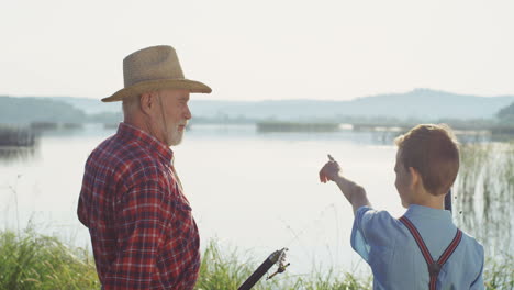 Rear-view-of-a-grandfather-and-grandson-holding-their-fishings-rods-and-talking-on-the-shore-of-the-river