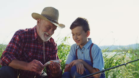 Old-grandpa-and-grandson-sitting-on-the-river-bank-and-looking-at-the-fish-they-caught