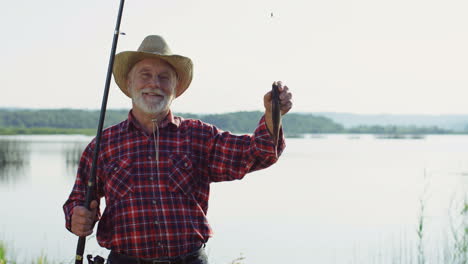 Senior-fisherman-in-a-hat-posing-for-the-camera-and-smiling-with-a-fish-on-a-rod-on-the-shore-of-a-lake-in-the-morning