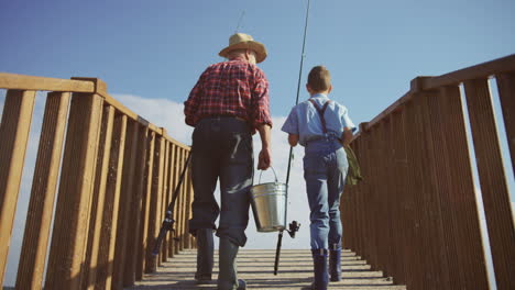 Rear-view-of-a-senior-man-and-his-small-grandson-walking-on-the-wooden-bridge-with-equipment-for-fishing