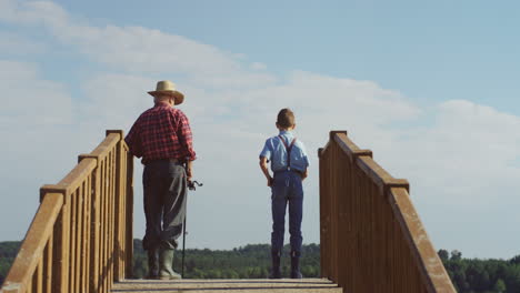 Rear-view-of-a-teen-boy-and-his-grandfather-fishing-in-the-wooden-bridge