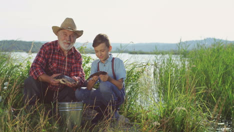 Smiling-old-grandpa-and-his-grandson-looking-at-the-camera-holding-fishes-in-the-shore-of-a-lake