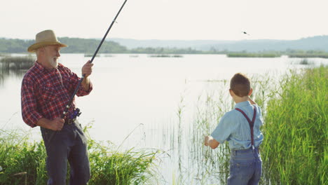Senior-caucasian-man-in-a-hat-catching-a-fish-from-the-lake-with-a-rod-and-his-little-son-taking-the-fish-off-the-hook