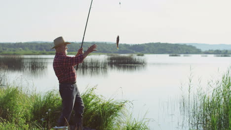 Viejo-Pescador-Caucásico-De-Pelo-Gris-Con-Un-Sombrero-Atrapando-Un-Pez-Del-Lago-Con-Una-Caña