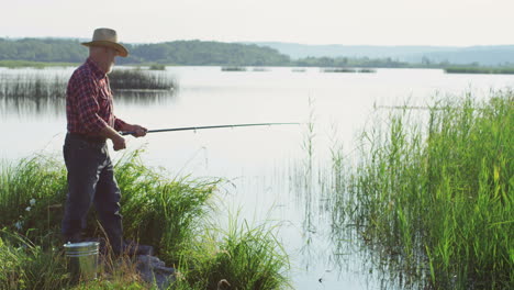 Side-view-of-a-old-man-in-a-hat-standing-on-the-lake-shore-and-fishing-with-a-rod