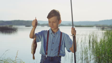 Cute-teenage-boy-posing-for-the-camera-with-a-fish-on-a-rod-while-standing-at-the-lake-shore-in-the-morning
