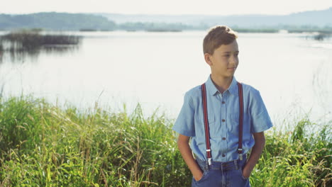 Portrait-of-the-cute-small-teen-boy-standing-on-the-big-lake-shore-and-looking-at-the-camera