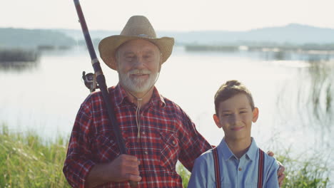 Portrait-of-a-gray-haired-fisherman-with-his-grandson-standing-at-the-lake-shore,-looking-at-each-other-and-smiling-to-the-camera