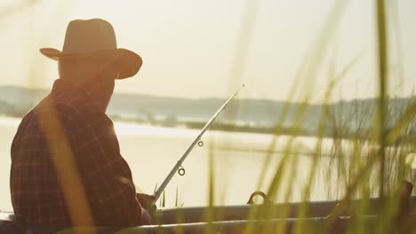 Side-view-of-a-senior-Caucasian-man-in-a-hat-catching-a-fish-in-a-boat-at-the-lake-in-the-morning