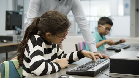 Focused-girl-in-eyeglasses-sitting-and-doing-task-on-computer-while-unrecognizable-teacher-looking-at-her