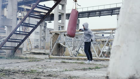 Caucasian-man-in-sportswear-hitting-a-punching-bag-outdoors-an-abandoned-factory-on-a-cloudy-morning