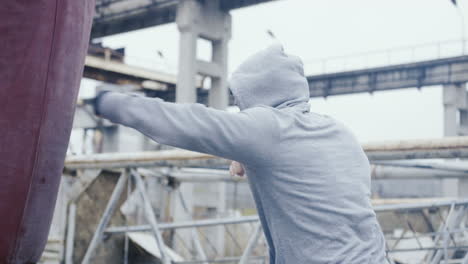 Side-view-of-caucasian-man-in-sportswear-hitting-a-punching-bag-outdoors-an-abandoned-factory-on-a-cloudy-morning