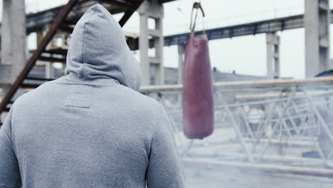Rear-view-of-caucasian-man-in-sportswear-hitting-a-punching-bag-outdoors-an-abandoned-factory-on-a-cloudy-morning