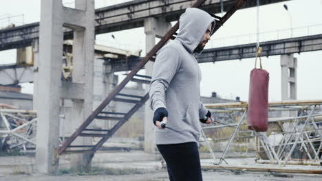 Caucasian-man-with-beard-in-a-grey-hoodie-jumping-rope-outdoors-an-abandoned-factory-on-a-cloudy-morning