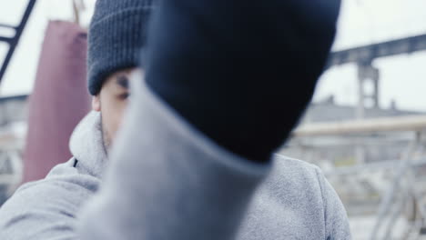 Close-up-view-of-handsome-bearded-caucasian-man-in-grey-hoodie-and-boxing-to-the-camera-outdoors-an-abandoned-factory-on-a-cloudy-morning