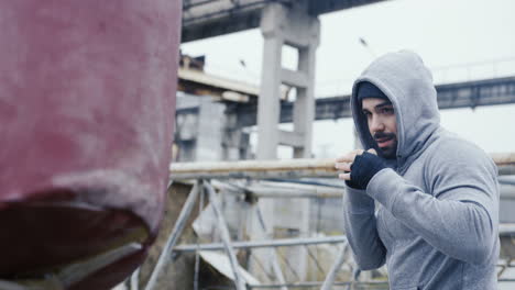 Side-view-of-caucasian-man-in-sportswear-hitting-a-punching-bag-outdoors-an-abandoned-factory-on-a-cloudy-morning