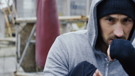 Close-up-view-of-handsome-bearded-caucasian-man-wearing-grey-hoodie-and-boxing-to-the-camera-outdoors-an-abandoned-factory-on-a-cloudy-morning