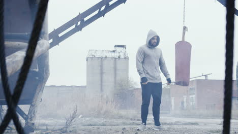 Caucasian-man-wearing-grey-hoodie-and-finising-his-boxing-training-near-the-red-punching-bag-outdoors-an-abandoned-factory-on-a-cloudy-morning