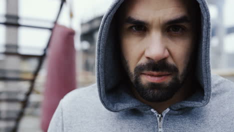 Close-up-view-of-handsome-bearded-man-with-grey-hoodie-and-looking-with-serious-expression-at-the-camera-outdoors-an-abandoned-factory-on-a-cloudy-morning