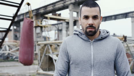 Handsome-bearded-sportsman-looking-with-serious-expression-at-the-camera-outdoors-an-abandoned-factory-on-a-cloudy-morning