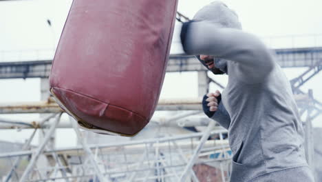 Side-view-of-caucasian-man-in-grey-hoodie-hitting-a-punching-bag-outdoors-an-abandoned-factory-on-a-cloudy-morning