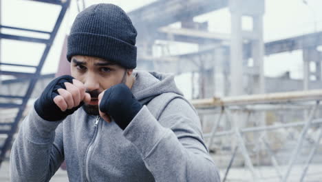 Handsome-bearded-caucasian-man-boxing-to-the-camera-outdoors-an-abandoned-factory-on-a-cloudy-morning