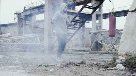Close-up-view-of-caucasian-man-with-beard-in-a-grey-hoodie-jumping-rope-outdoors-an-abandoned-factory-on-a-cloudy-morning