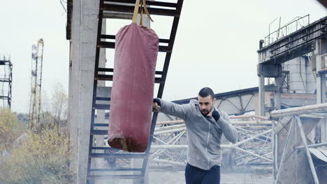 Caucasian-man-in-grey-hoodie-hitting-a-punching-bag-outdoors-an-abandoned-factory-on-a-cloudy-morning