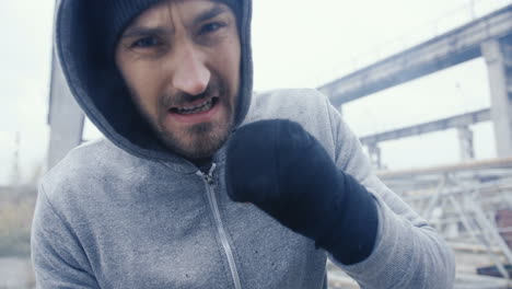 Caucasian-sportsman-in-grey-hoodie-boxing-to-the-camera-outdoors-an-abandoned-factory-on-a-cloudy-morning