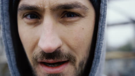 Close-up-view-of-the-sweaty-face-of-the-male-boxer-in-grey-hoodie-during-a-training-outdoors-an-abandoned-factory-on-a-cloudy-morning