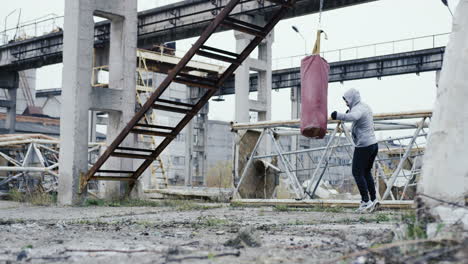 Distant-view-of-caucasian-man-in-grey-hoodie-hitting-a-punching-bag-outdoors-an-abandoned-factory-on-a-cloudy-morning