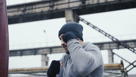 Close-up-view-of-caucasian-man-in-grey-beanie-and-sportswear-hitting-a-punching-bag-outdoors-an-abandoned-factory-on-a-cloudy-morning