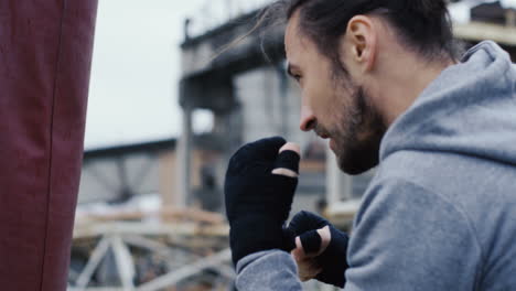 Close-up-view-of-caucasian-man-in-sportswear-hitting-a-punching-bag-outdoors-an-abandoned-factory-on-a-cloudy-morning
