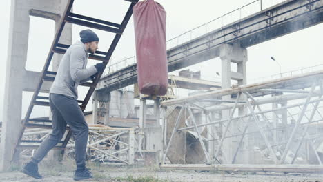 Caucasian-man-in-grey-beanie-and-sportswear-hitting-a-punching-bag-outdoors-an-abandoned-factory-on-a-cloudy-morning