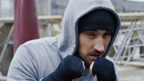 Close-up-view-of-caucasian-bearded-caucasian-man-in-grey-hoodie-and-boxing-to-the-camera-outdoors-an-abandoned-factory-on-a-cloudy-morning