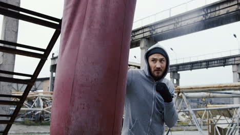 Side-view-of-caucasian-bearded-caucasian-man-in-grey-hoodie-hitting-a-punching-bag-outdoors-an-abandoned-factory-on-a-cloudy-morning