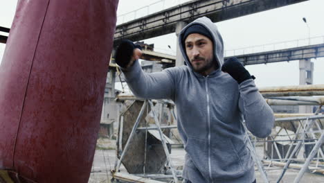 Side-view-of-caucasian-bearded-caucasian-man-in-grey-hoodie-hitting-a-punching-bag-outdoors-an-abandoned-factory-on-a-cloudy-morning