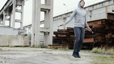 Caucasian-sportsman-with-beard-in-a-grey-hoodie-jumping-rope-outdoors-an-abandoned-factory-on-a-cloudy-morning