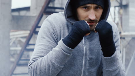 Caucasian-bearded-caucasian-man-in-grey-hoodie-and-boxing-to-the-camera-outdoors-an-abandoned-factory-on-a-cloudy-morning