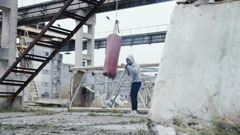 Distant-view-of-caucasian-man-in-grey-hoodie-hitting-a-punching-bag-outdoors-an-abandoned-factory-on-a-cloudy-morning