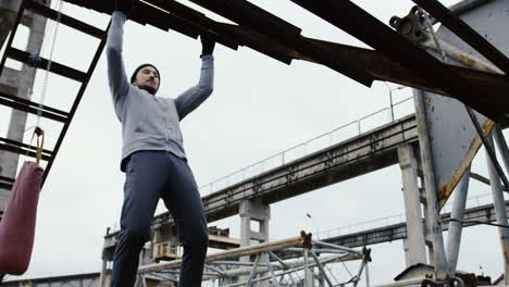 Young-handsome-sportsman-doing-pull-ups-on-the-metal-construction-in-the-ruins-of-an-abandoned-factory-on-a-cloudy-morning
