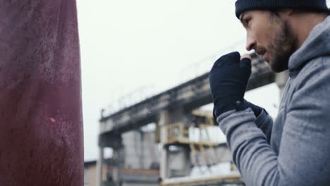 Side-view-of-caucasian-man-in-black-beanie-hitting-a-punching-bag-outdoors-an-abandoned-factory-on-a-cloudy-morning