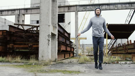 Distant-view-of-caucasian-sportsman-with-beard-in-a-grey-hoodie-jumping-rope-outdoors-an-abandoned-factory-on-a-cloudy-morning
