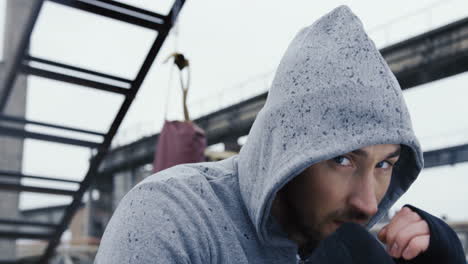 Close-up-view-of-handsome-bearded-caucasian-man-in-grey-hoodie-and-boxing-to-the-camera-outdoors-an-abandoned-factory-on-a-cloudy-morning