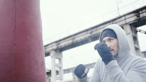 Close-up-view-of-caucasian-man-in-grey-hoodie-hitting-a-punching-bag-outdoors-an-abandoned-factory-on-a-cloudy-morning