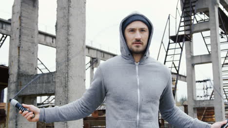 Portrait-of-caucasian-sportsman-with-beard-in-a-grey-hoodie-jumping-rope-outdoors-an-abandoned-factory-on-a-cloudy-morning